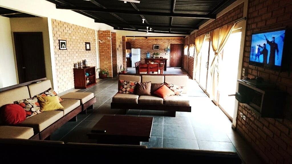 An open living area is highlighted by comfortable seating with colorful cushions. Large windows allow natural light to fill the space, while a dining area with a wooden table is visible in the background. A flat-screen TV is mounted on the wall.