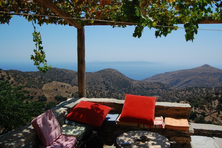 A sheltered outdoor seating area features comfortable cushions in various colors, arranged around a small table. Overhead, grapevines provide shade, while a panoramic view of rolling hills and the Aegean Sea is visible in the background, enhancing the peaceful setting.
