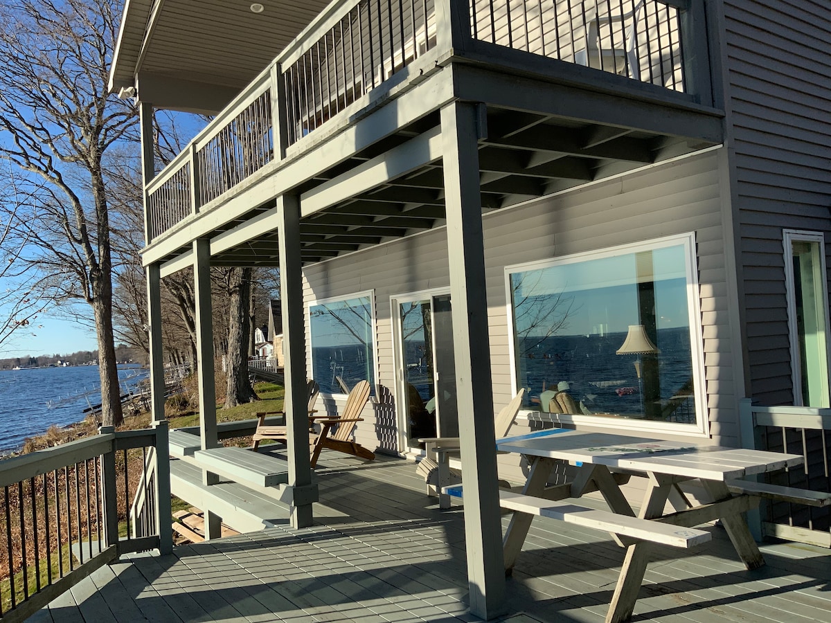 A multi-level deck extends along the house, featuring several picnic-style tables and lounge chairs arranged for relaxation. Large windows offer views of the lake, allowing natural light to illuminate the space. The tranquil water and surrounding trees create a peaceful backdrop.