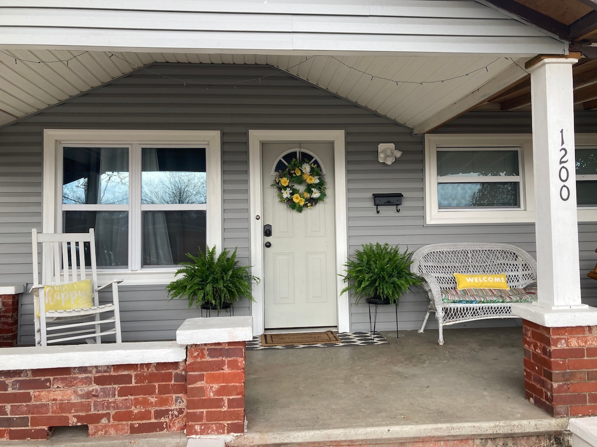 The front porch features a welcoming entryway with a neutral door surrounded by light-colored trim. A floral wreath adorns the door, while potted greenery flanks either side. A rocking chair and a wicker bench provide seating under the covered space.