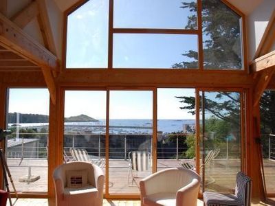 A spacious living area features large glass windows that provide a panoramic view of the sea and nearby islands. Comfortable seating is arranged below the windows, inviting relaxation while taking in the scenic landscape. Natural light fills the space, enhancing the warm wooden interior.