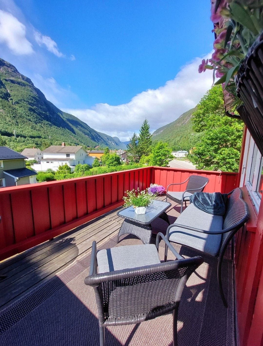 A red balcony overlooks a serene landscape, featuring comfortable seating with cushions and a low table. Potted flowers add a touch of colour. Scenic mountains and greenery can be seen in the background, creating a relaxing outdoor space.
