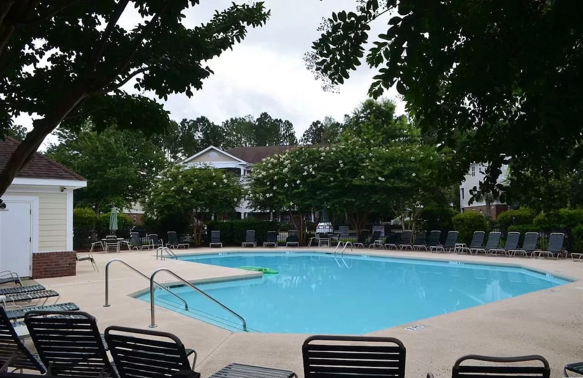 A swimming pool is surrounded by green trees and lounge chairs, offering space for relaxation. The pool features a shallow entry with handrails for easy access. Townhouse buildings are visible in the background, providing a serene setting within the community.