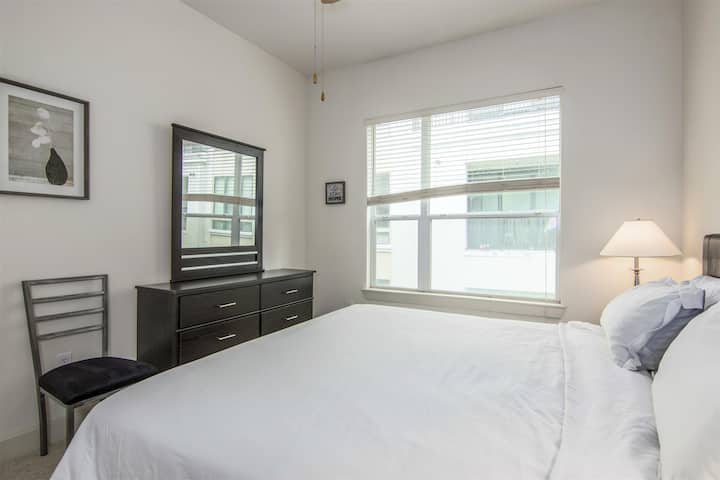 bedroom featuring a large window with blinds, a comfortable bed with white linens, and a dark wood dresser with a matching mirror.