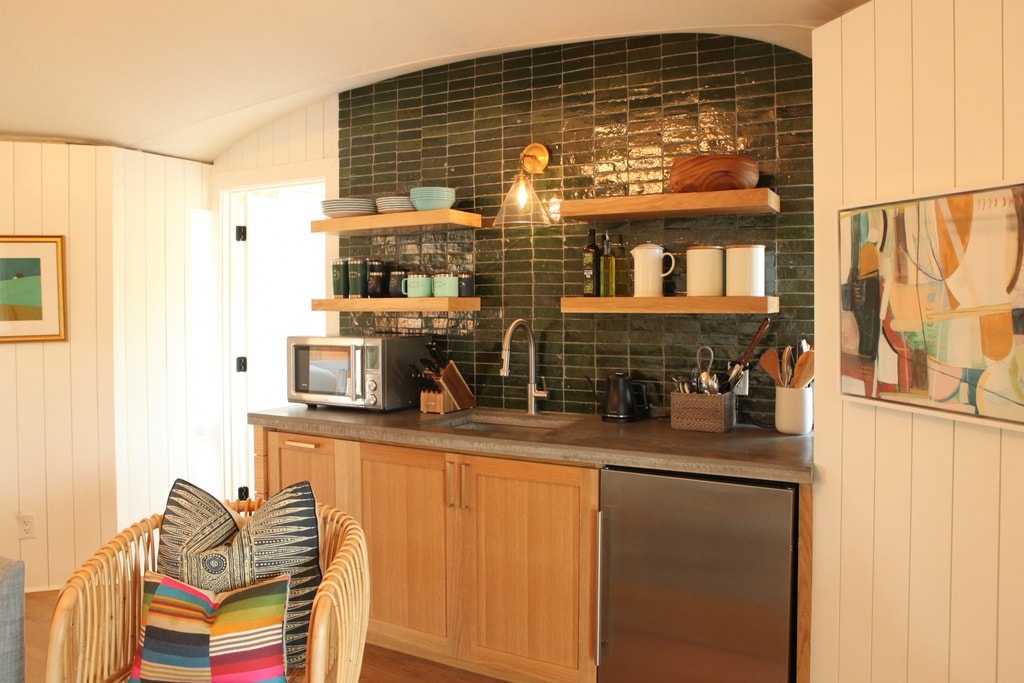 A well-equipped kitchenette features wooden shelves with bowls and utensils arranged above a countertop. A microwave and a stainless steel refrigerator are present, along with a sink. Colorful cushions are displayed on a nearby wicker chair, contributing to a relaxed atmosphere.