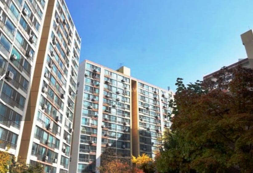 The image captures the exterior of a tall apartment building against a clear blue sky. The structure features multiple floors of large windows, showcasing a modern architectural style with a blend of concrete and glass. Trees with autumn foliage are partially visible in the foreground.