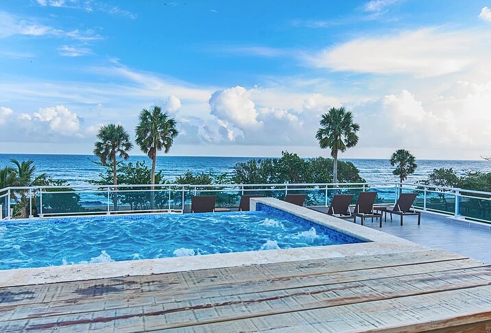 A serene view of the ocean is visible from the beachfront rooftop terrace. The infinity pool and a smaller whirlpool are situated beside a wooden deck, which is lined with sun loungers. Palm trees frame the scenic coastline under a bright, blue sky.