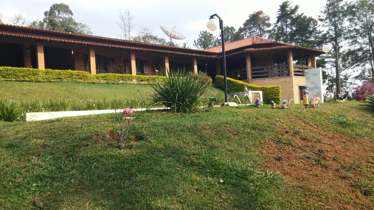 The exterior of a spacious house is visible, set against a lush green hillside. Flower beds with various plants are arranged along the pathway. Surrounding the house are manicured lawns and tall trees, contributing to a serene atmosphere.