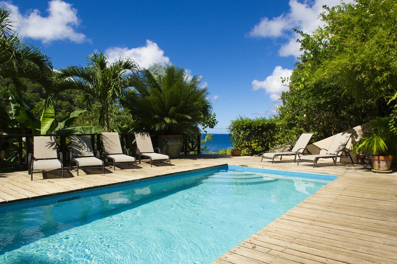 A serene pool area features a clear blue pool surrounded by wooden decking. Sun loungers are positioned on one side, with lush greenery and flowering plants adding to the natural beauty. The ocean can be seen in the distance under a bright blue sky with scattered clouds.