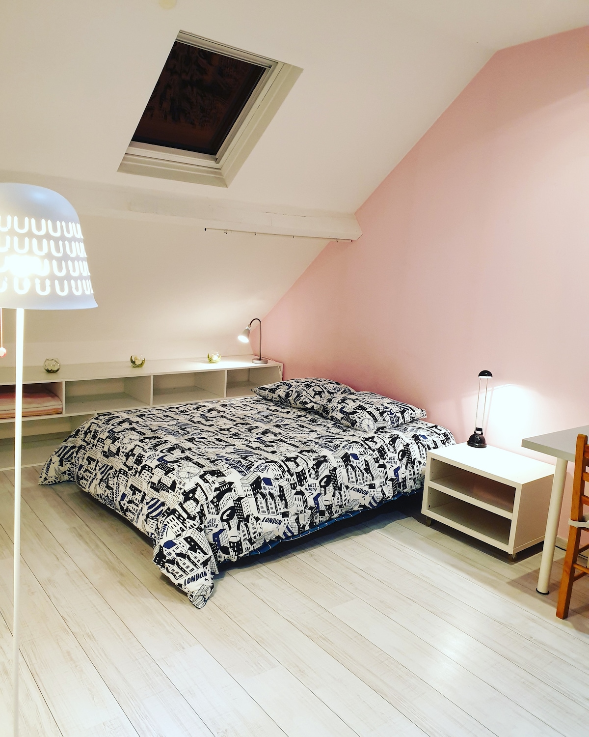 A spacious bedroom is presented with a double bed covered by a patterned comforter. A desk and lamp are situated next to the bed, with minimalist shelving along the wall. Natural light enters through a skylight, complementing soft pink walls.
