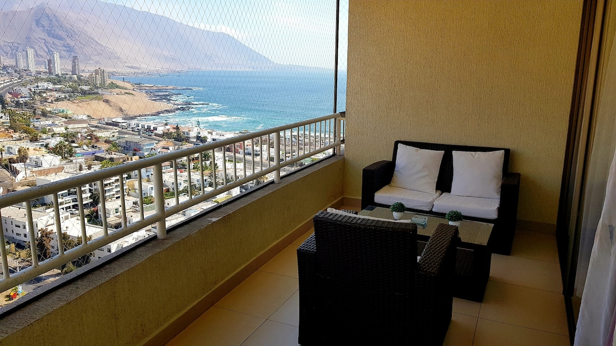 A comfortable terrace setup features outdoor seating with soft white cushions and a glass coffee table. The railing is protected by mesh for safety. A panoramic view of the coastline and mountains is visible in the background, offering a sense of openness and tranquility.