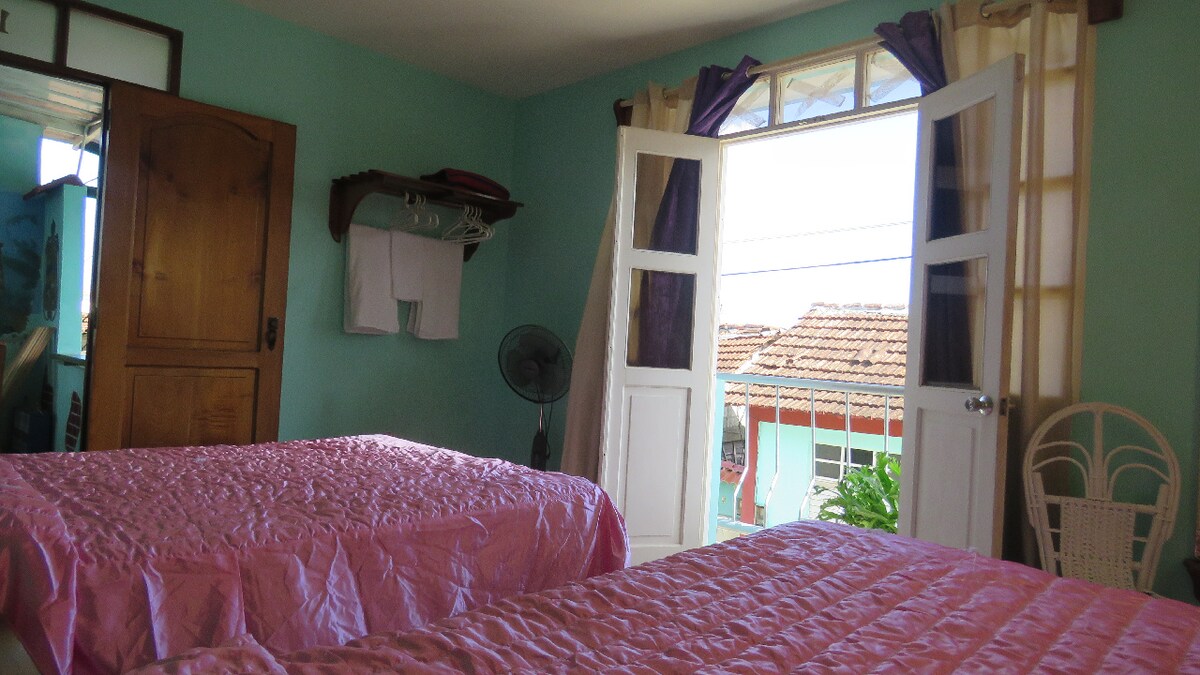 A bright room features two spacious beds covered with pink bedspreads. Light enters through a wide window flanked by curtains, creating a cheerful atmosphere. A wooden wardrobe is positioned against the wall, while a ceiling fan adds comfort to the space.