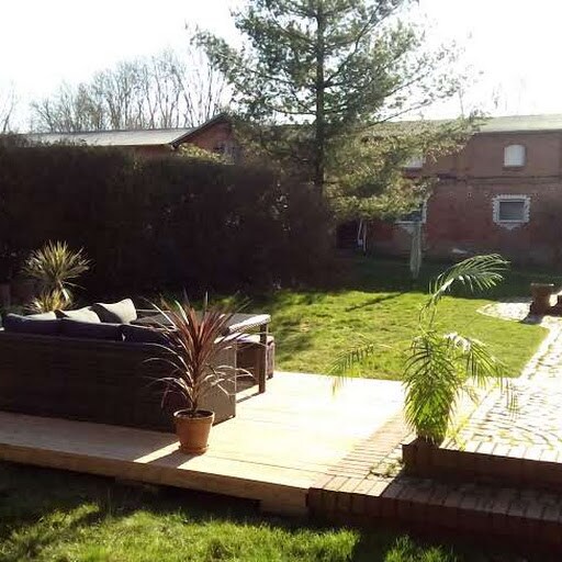 A sunlit terrace features a comfortable seating area with dark cushions surrounded by greenery. Potted plants add a touch of nature, while a spacious lawn invites relaxation and outdoor gatherings. The backdrop includes trees and rustic buildings, contributing to a serene environment.
