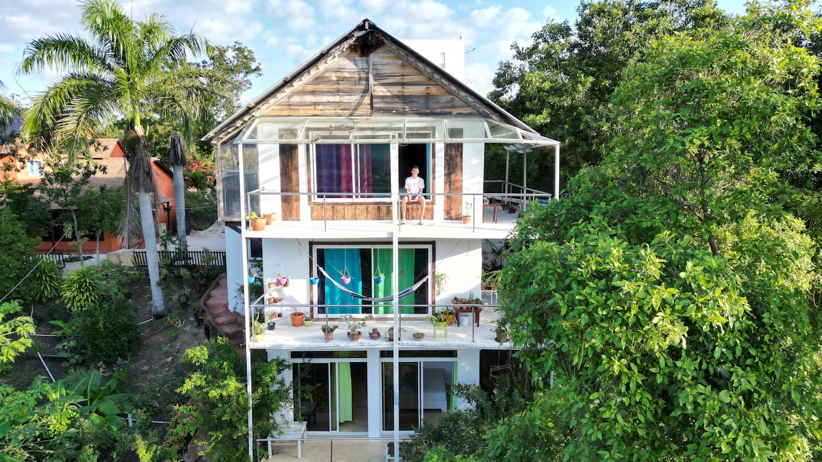 A two-story guest house is surrounded by lush greenery, showcasing a modern design with an open balcony on each level. Colorful curtains hang in the large windows, while potted plants add natural beauty to the outdoor spaces.