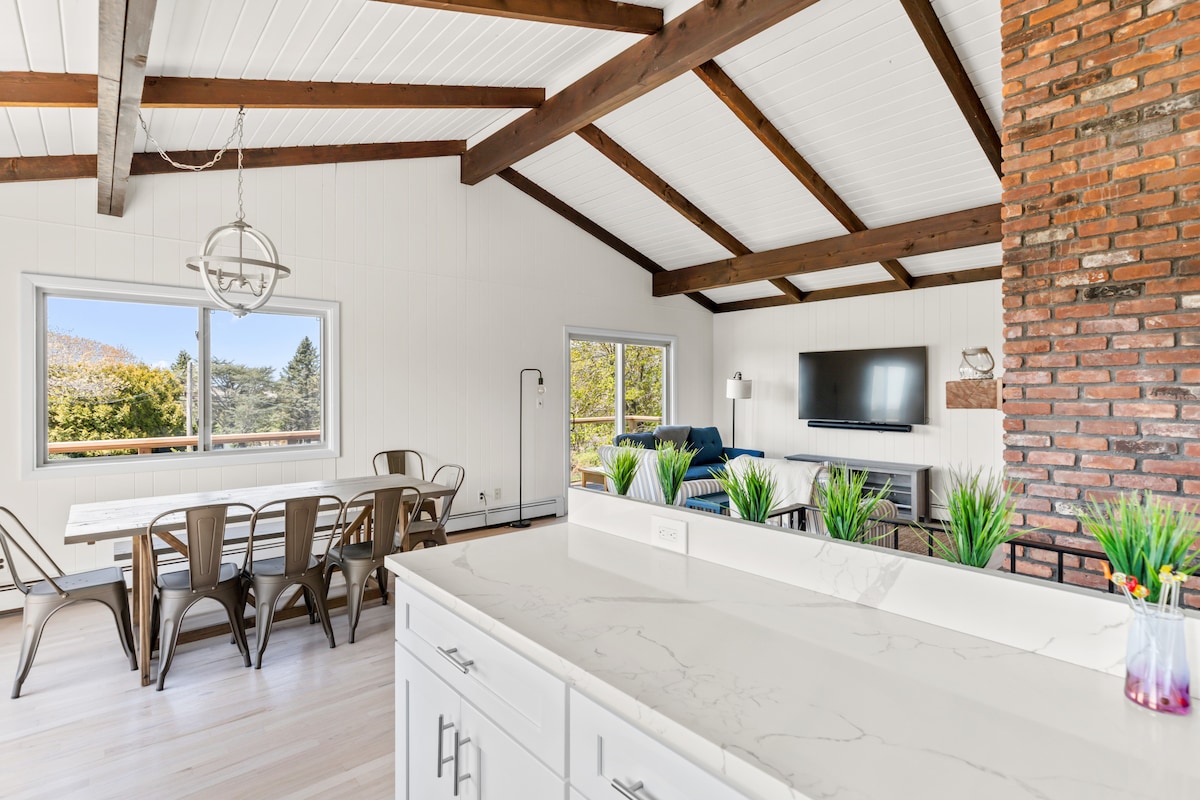 An open-concept space features a modern kitchen with white cabinetry and a marble countertop. A dining area with metal chairs overlooks large windows, showcasing outdoor views. Exposed wooden beams and a brick accent wall add character, while a comfortable living area is anchored by a wall-mounted television.