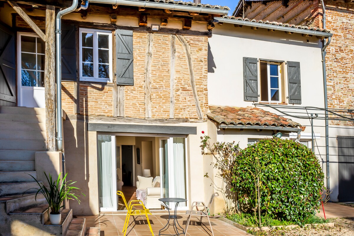 The exterior of the building showcases a blend of textured brick and wooden accents, framed by shuttered windows. A small patio area is visible, featuring a round table and two yellow chairs, complemented by greenery and natural stone. Soft sunlight bathes the space.