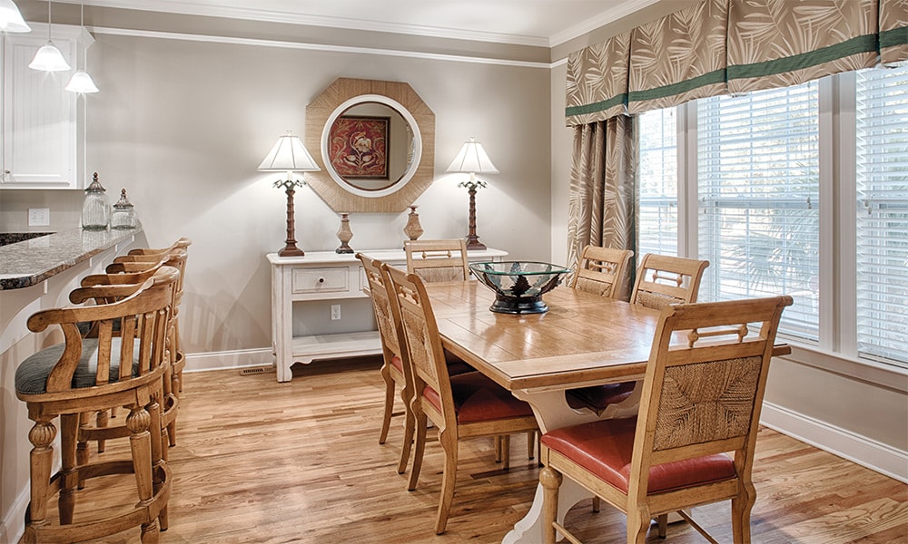 A dining area features a wooden table surrounded by six chairs with cushioned seats. Two lamps provide warm lighting atop side tables. A bowl sits in the center of the table, and large windows are adorned with patterned curtains, allowing natural light to fill the space.