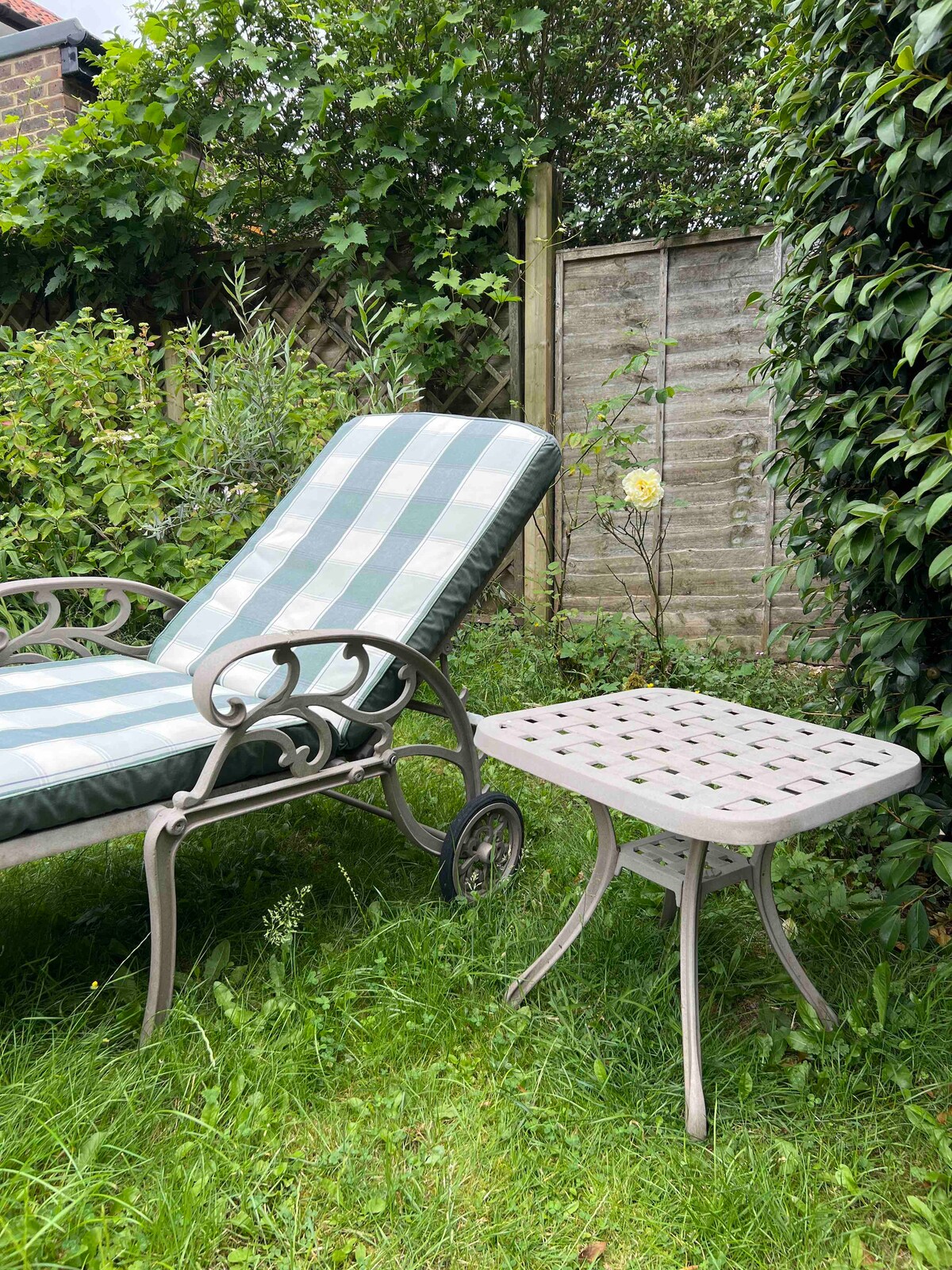 A lounge chair with a green and white checkered cushion sits beside a small, light-colored side table. The green lawn is surrounded by lush foliage, creating a serene garden space. A single yellow flower is visible among the greenery, enhancing the natural setting.