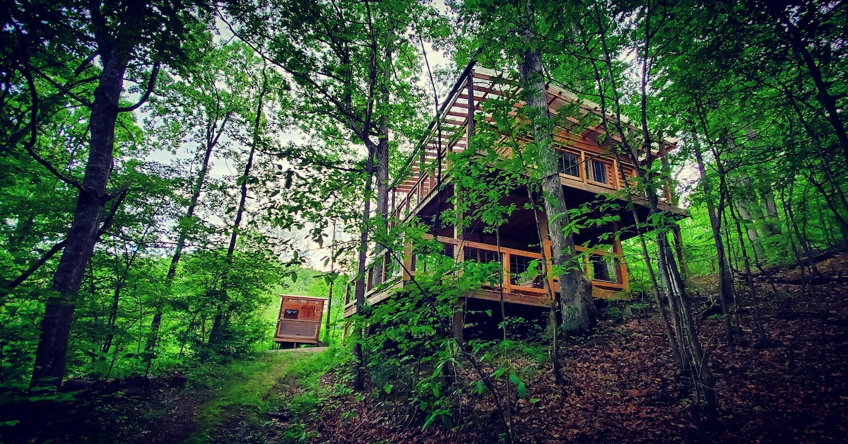 The Treetop Flyer has 4 full-sized beds (2 on the main floor and 2 in the loft). Wrap around porch with seating and deck below with swing, picnic table, and propane grill. Outhouse and firepit nearby.