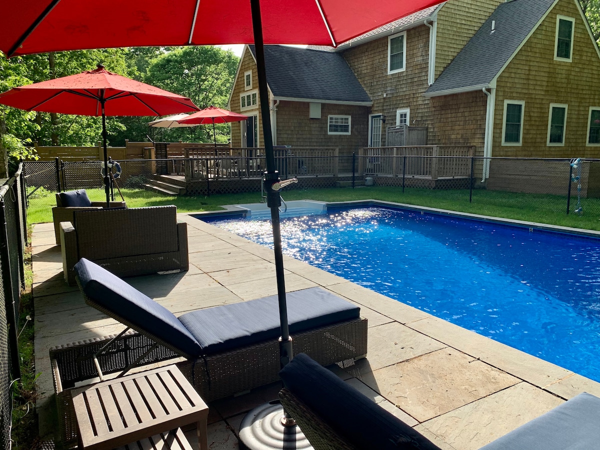 The outdoor area features a private, heated pool surrounded by a spacious patio. Comfortable lounge chairs with cushions are positioned under red umbrellas, providing shade. The fenced backyard is complemented by lush greenery and a wooden deck area visible in the background.
