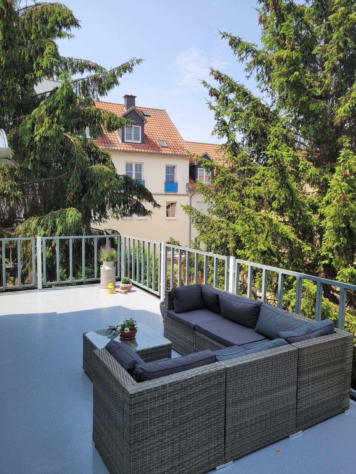 A private terrace features a seating area with a dark wicker sofa and a matching coffee table. Surrounding greenery provides a natural backdrop, while nearby buildings are partially visible in the background. The sky is clear, allowing for plenty of natural light.