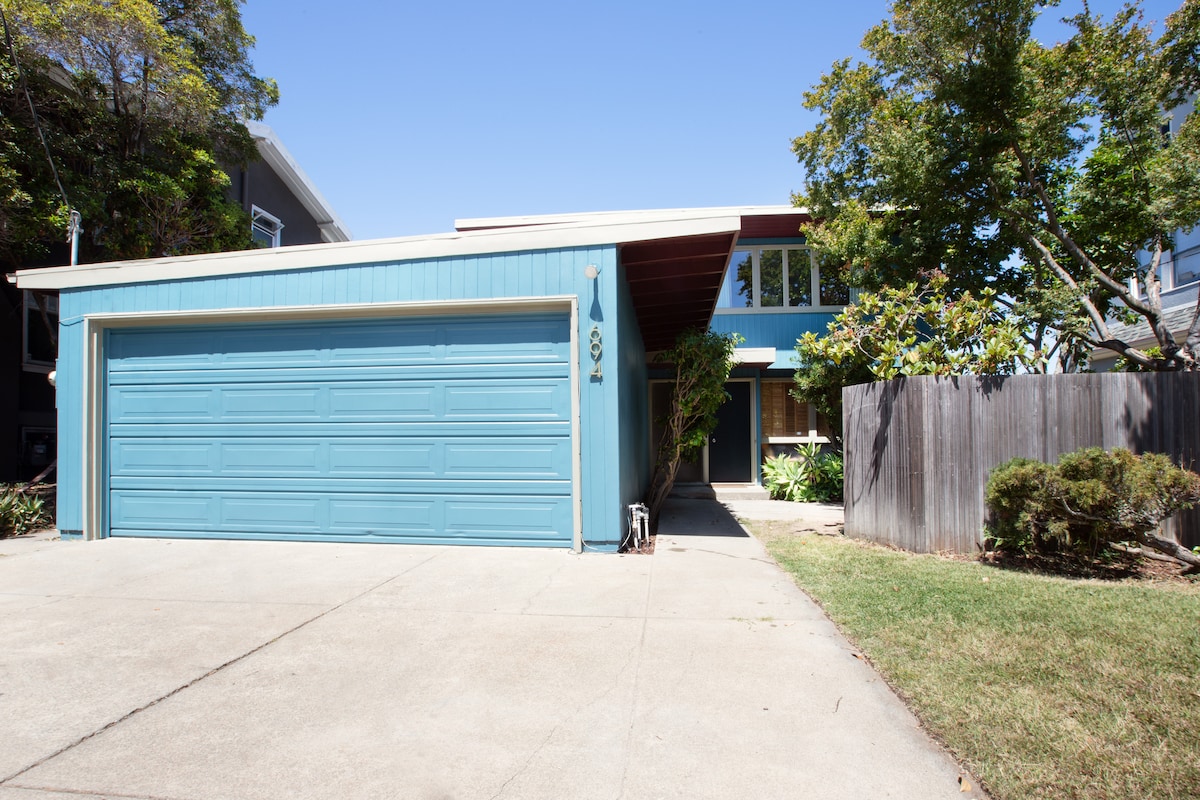 The exterior of the house is showcased with a blue facade and a two-car garage. A paved driveway leads to the entrance, bordered by well-maintained grass and shrubs. Large windows are visible, allowing natural light into the home.
