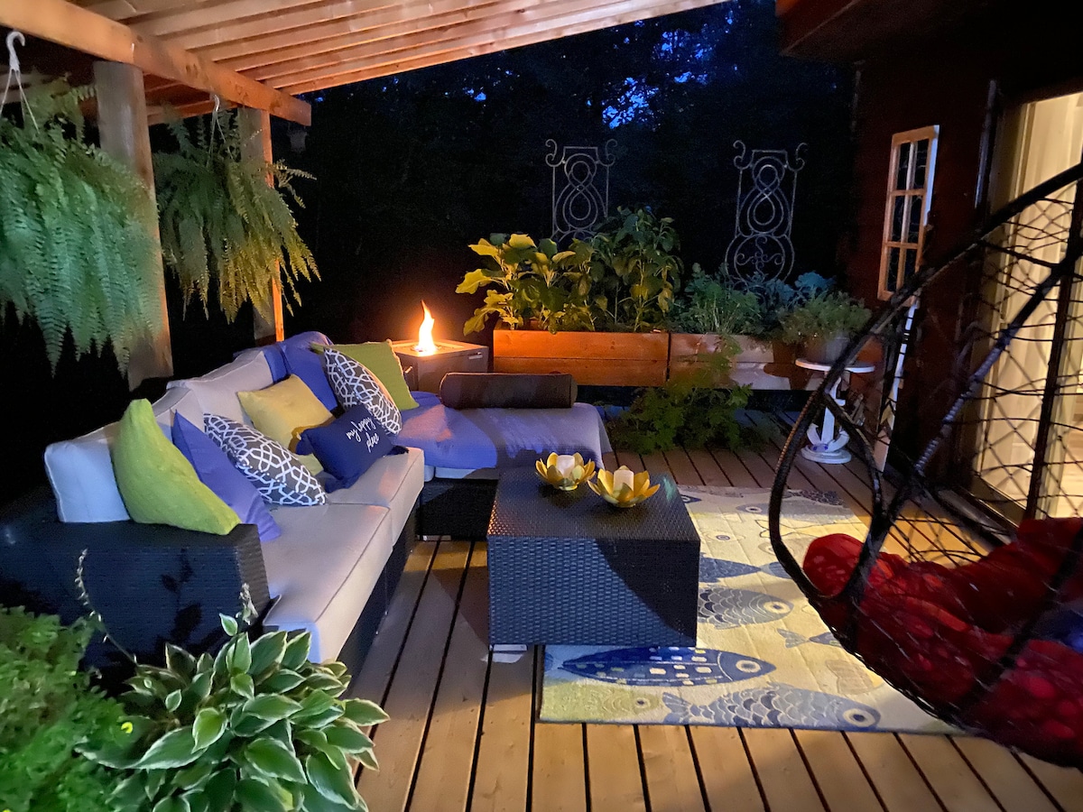 A screened porch is illuminated by soft lighting, featuring an outdoor sofa adorned with colorful cushions and a large swing chair. A propane fireplace provides warmth, and potted plants enhance the natural ambiance, creating a relaxing space for enjoying the evening.