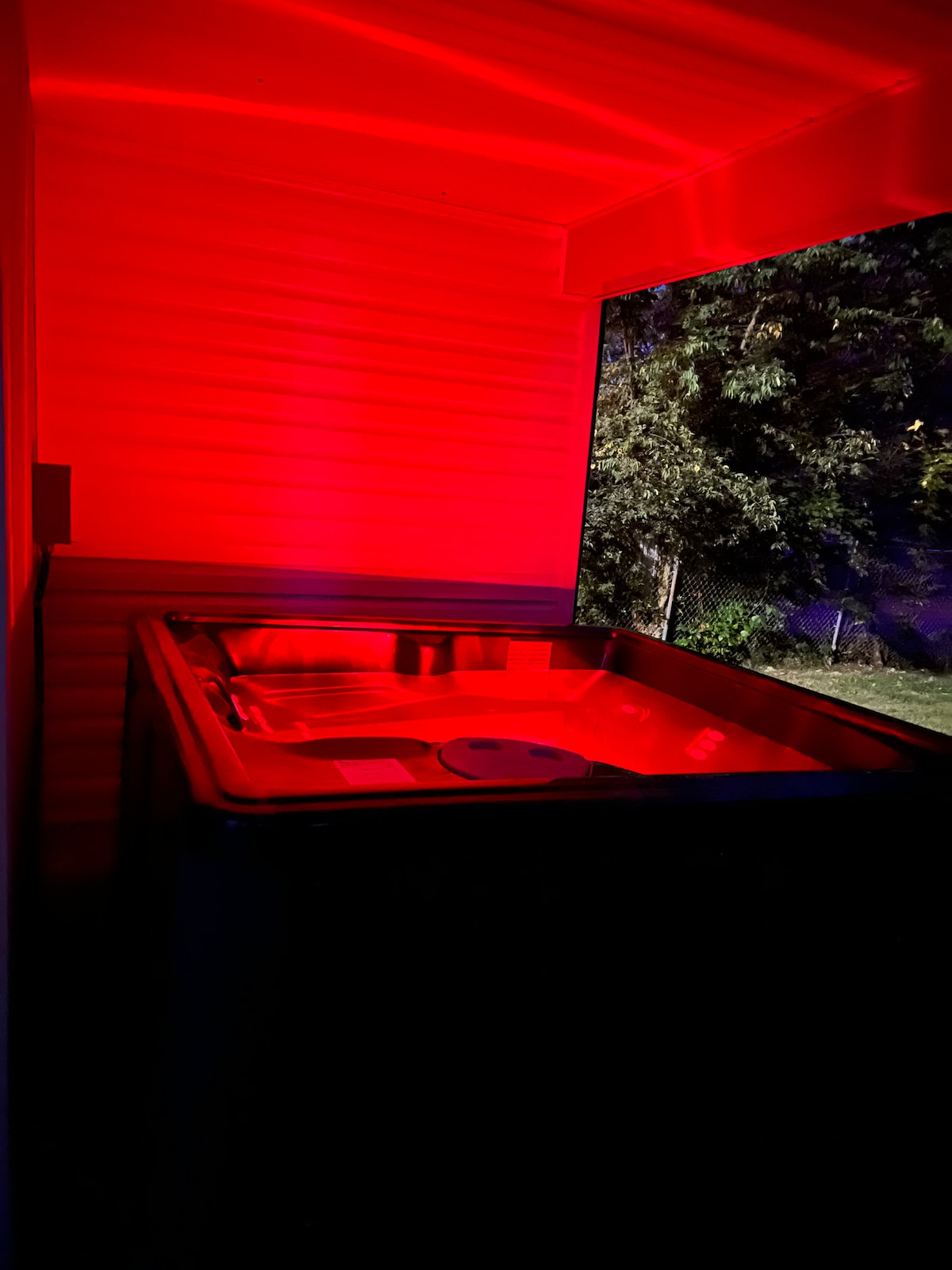 A hot tub is situated on a covered patio, featuring soft red lighting and surrounded by trees. The ambiance is enhanced by evening shadows, creating a serene outdoor space for relaxation.