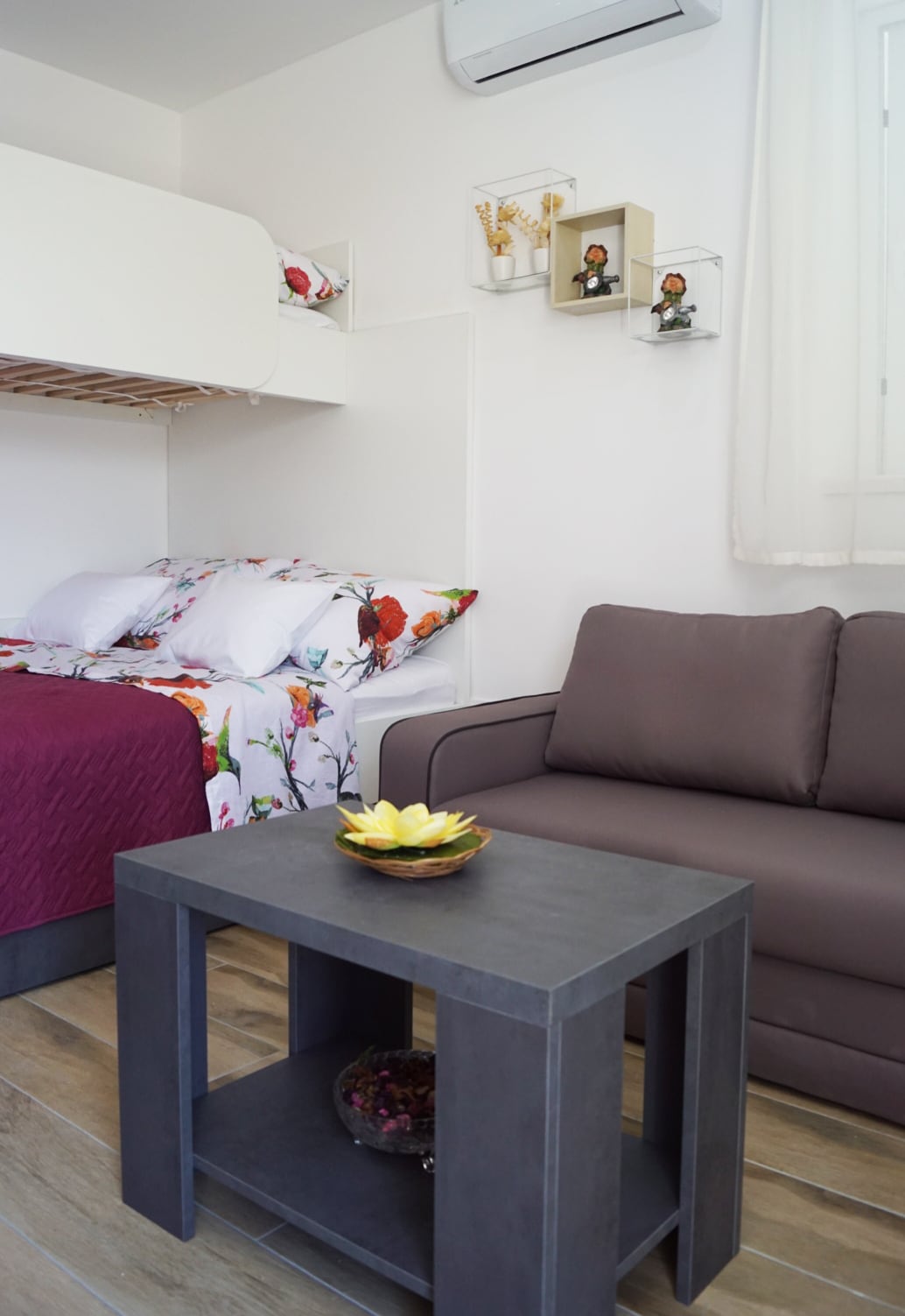 A studio room features a king-size bed adorned with floral bedding, complemented by a single bunk bed above. A gray sofa is positioned adjacent to a small table with a decorative bowl, enhancing the function and comfort of the space.