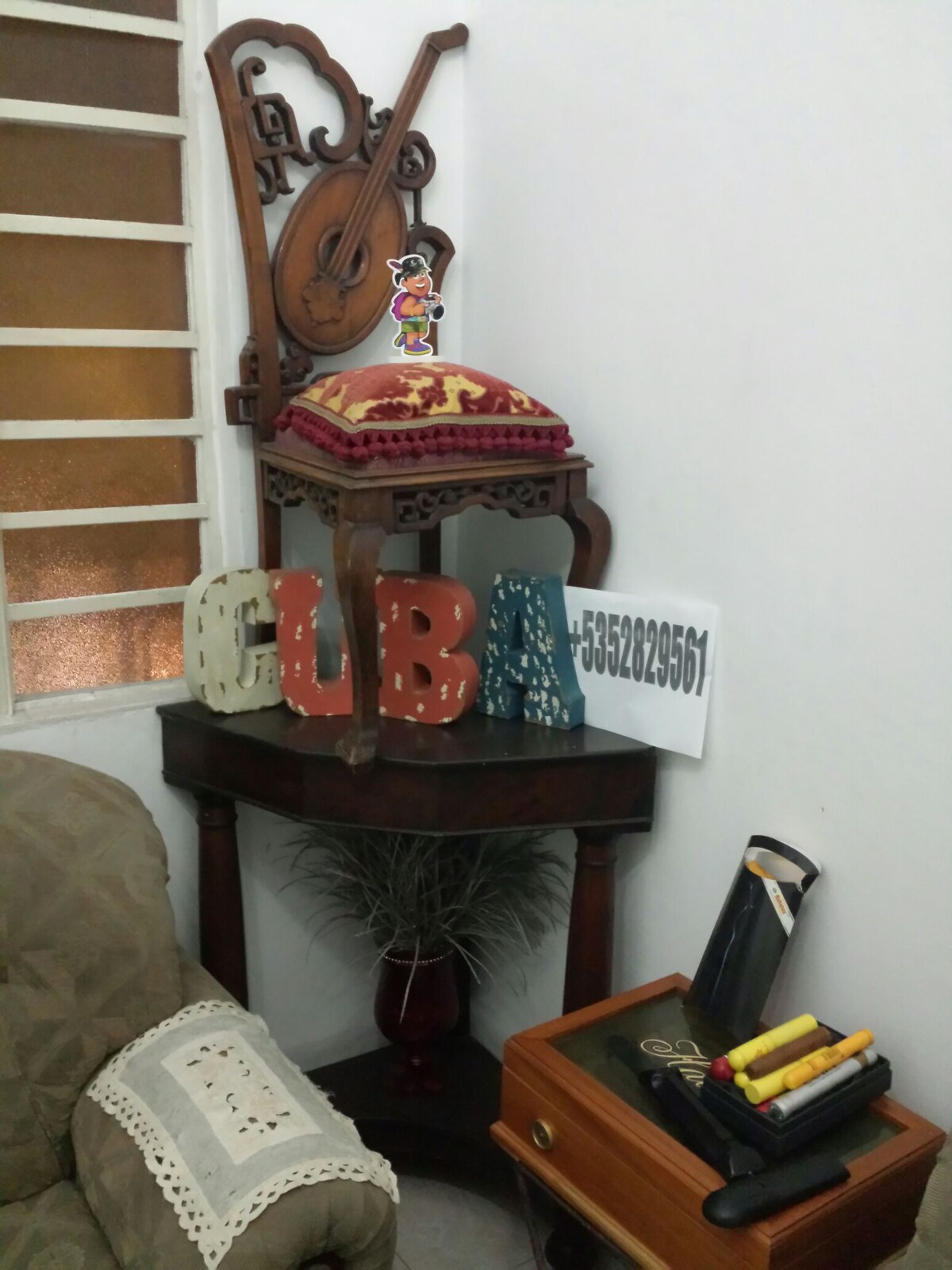 A decorative corner is displayed with a wooden chair featuring intricate carvings and a colorful cushion. The word 'CUBA' is formed with painted letters of varying sizes, resting on a small table adorned with a potted plant. A vintage-style box and art supplies complete the arrangement.