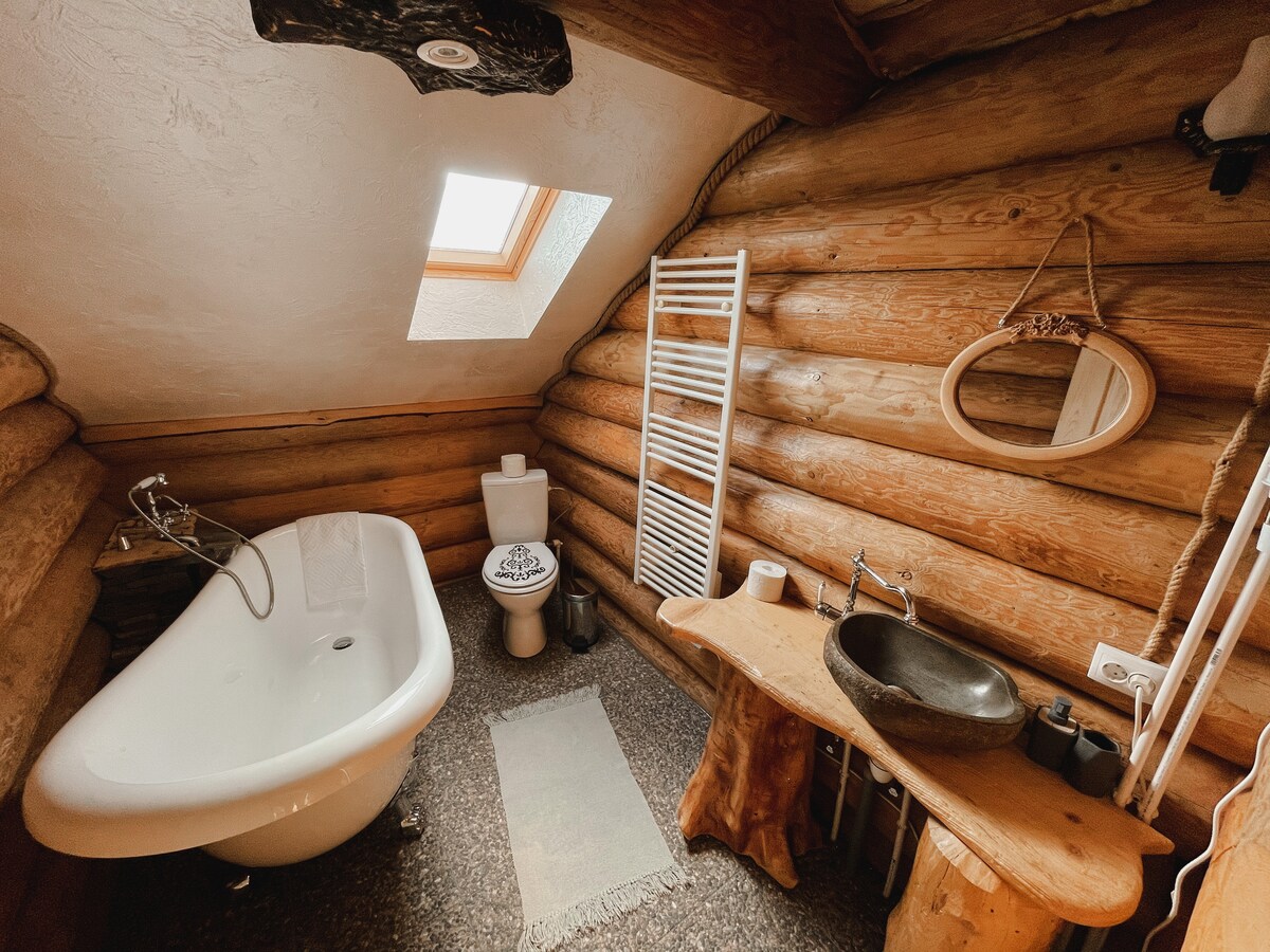 A rustic bathroom features a freestanding bathtub beneath a skylight, allowing natural light to fill the space. Wooden logs create the walls, while a unique wooden sink adds character. A small rug lies on the stone floor, and a towel radiator is visible alongside essential toiletries.