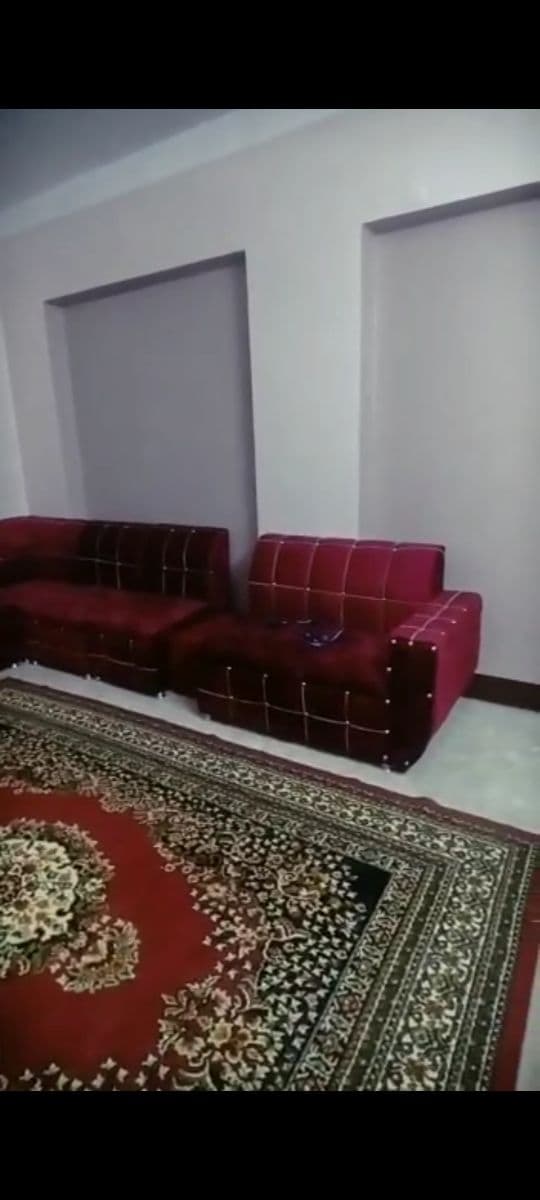 A seating area features deep red sofas with a grid pattern, positioned against light-colored walls. A decorative rug with intricate designs complements the furniture, creating a comfortable space. Soft lighting illuminates the area, enhancing the overall inviting appearance.
