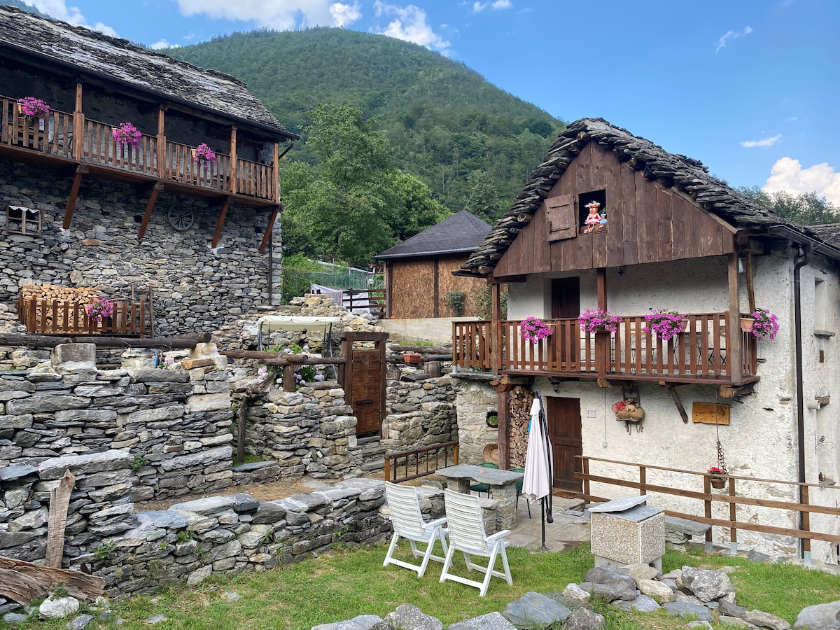 The exterior of the rustic home is surrounded by stone walls and features wooden balconies adorned with pink flowers. A green lawn provides space for outdoor seating arrangements, including chairs and a table, alongside a large stone structure that hints at historical significance.