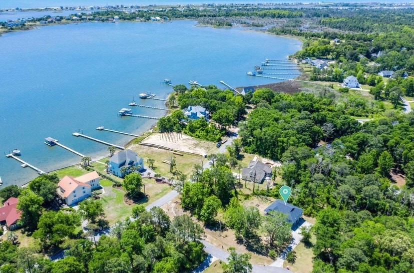 An aerial view showcases the coastal neighborhood and its proximity to the bay. Multiple docks are visible along the shoreline, surrounded by lush greenery and residential properties. The tranquil bay reflects the clear blue sky, emphasizing the scenic environment ideal for relaxation.