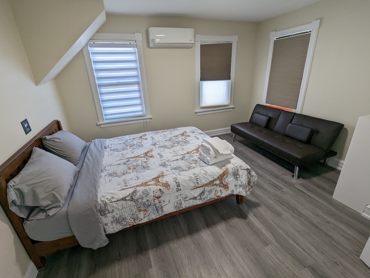 A well-lit bedroom showcases a wooden bed with a light-colored comforter adorned with a subtle design. A sleek, dark sofa provides additional seating, and two windows are fitted with light-filtering shades. An air conditioning unit is mounted on the wall.