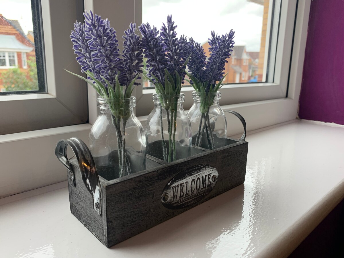 Four small glass bottles containing artificial lavender flowers are arranged in a black wooden tray. The tray has a 'WELCOME' sign attached, and natural light streams in from the window, highlighting the decorative element against the vibrant wall.