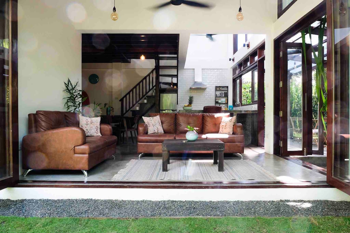 An open living area features a comfortable brown leather sofa set around a coffee table, with decorative pillows adding a touch of style. Large glass doors provide a view of the private garden, while a modern kitchen is visible in the background. Light fixtures hang above, enhancing the airy atmosphere.