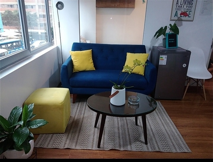 A comfortable living area is presented with a blue sofa adorned with yellow cushions. A round glass coffee table sits atop a textured rug, accompanied by a small ottoman. A potted plant adds a touch of greenery, while a fridge is positioned nearby.