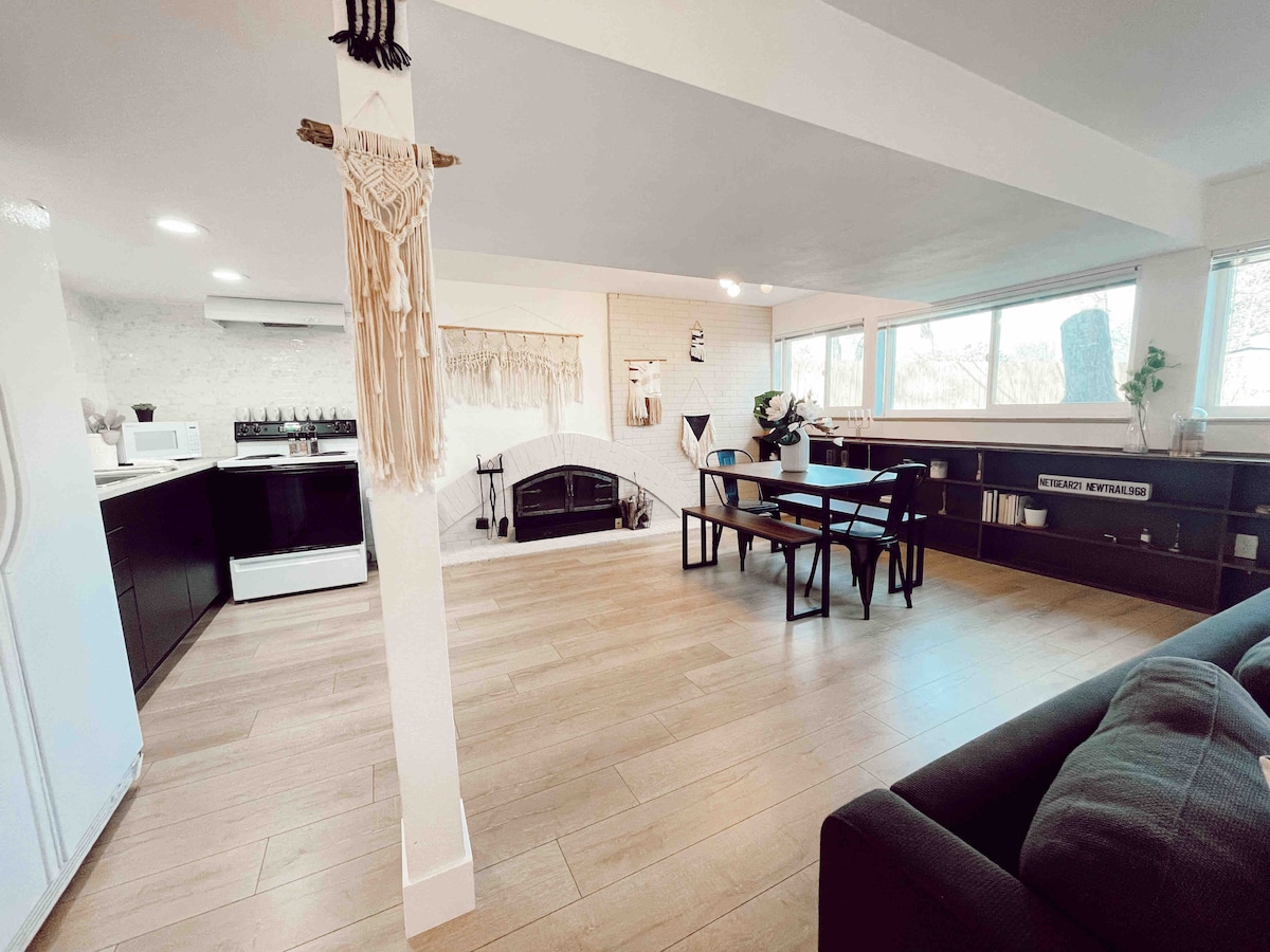 An open-concept living area features large windows allowing natural light to fill the space. A dining table with black chairs is positioned beside a cozy seating area, which includes a modern sectional sofa. A decorative wall hanging adds a touch of detail to the inviting atmosphere.
