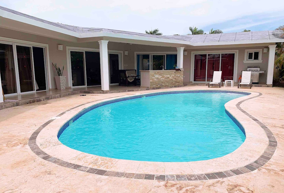 A private pool with a curved design is surrounded by a spacious patio, featuring lounge chairs for relaxation. The area is framed by large glass doors leading into an open living space, allowing for an airy and bright atmosphere.