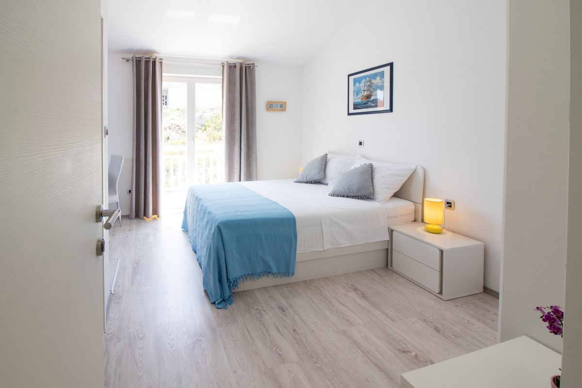 A bright sleeping area features a spacious bed dressed in white linens, complemented by two decorative pillows. Natural light enters through the window, framed by light gray curtains. A nightstand with a lamp sits alongside the bed, while a minimalist chair is positioned nearby.