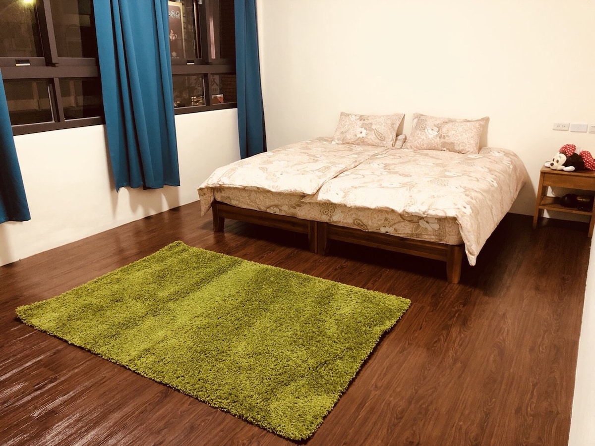 A spacious sleeping area features two beds with light-colored bedding positioned on wooden flooring. A plush green rug adds comfort underfoot, while large windows framed by blue curtains allow natural light to brighten the room.