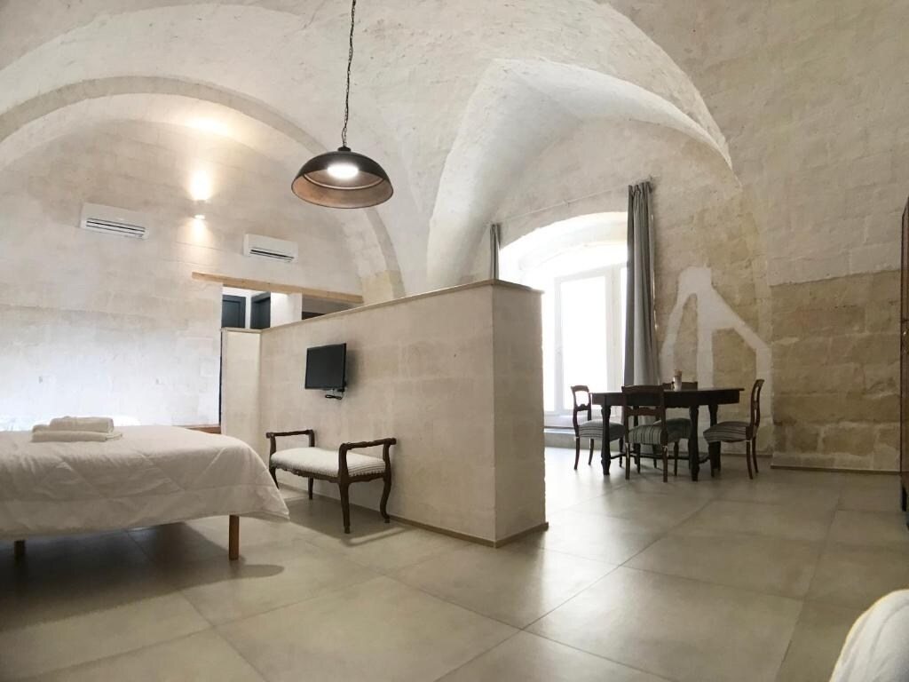 A spacious loft features vaulted ceilings with exposed brick, creating an airy atmosphere. A cozy sleeping area is positioned next to a small dining table. Natural light flows through large windows, and a wall-mounted TV is visible near the seating area.