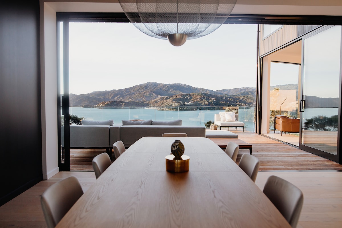 A spacious dining area features a large wooden table surrounded by comfortable chairs. Floor-to-ceiling glass doors open to reveal stunning views of the water and distant mountains, while natural light fills the space, enhancing the minimalist décor.