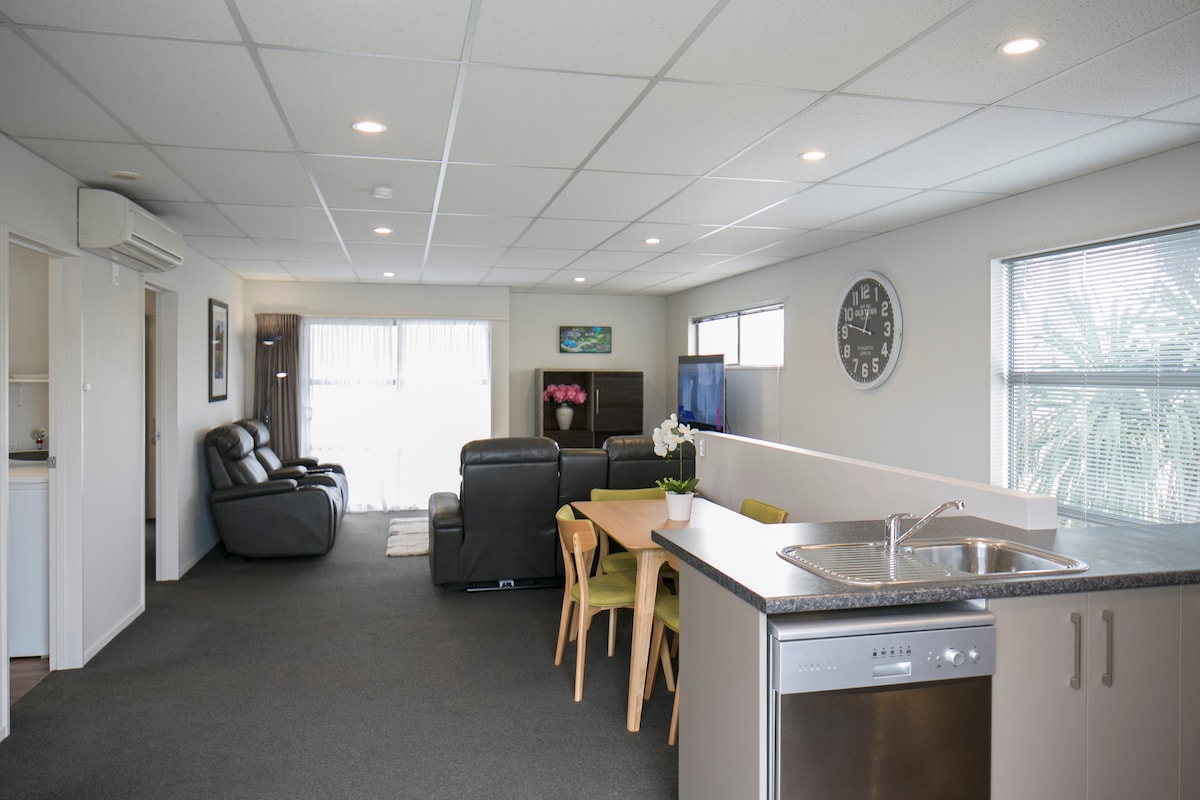 An open-plan living and dining area features a well-appointed kitchen with modern appliances. A dining table is set with wooden chairs nearby. Two comfortable recliners are positioned for relaxation, with natural light streaming in through large windows.