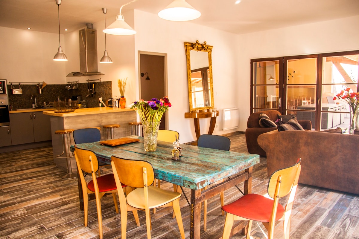 A spacious living area features a large wooden dining table surrounded by colorful chairs. A well-equipped kitchen is visible in the background, complemented by contemporary pendant lighting. Comfortable seating is provided by a plush brown sofa, enhancing a relaxed communal atmosphere.