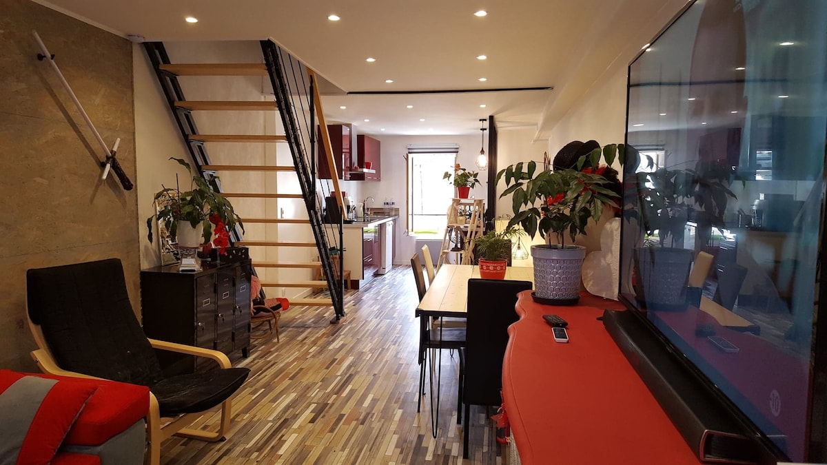 An inviting living space features wooden flooring and soft lighting. A cozy seating area includes a black lounge chair, while a dining table is set for multiple guests. A well-equipped kitchen is visible in the background, and several plants add a touch of greenery throughout the area.