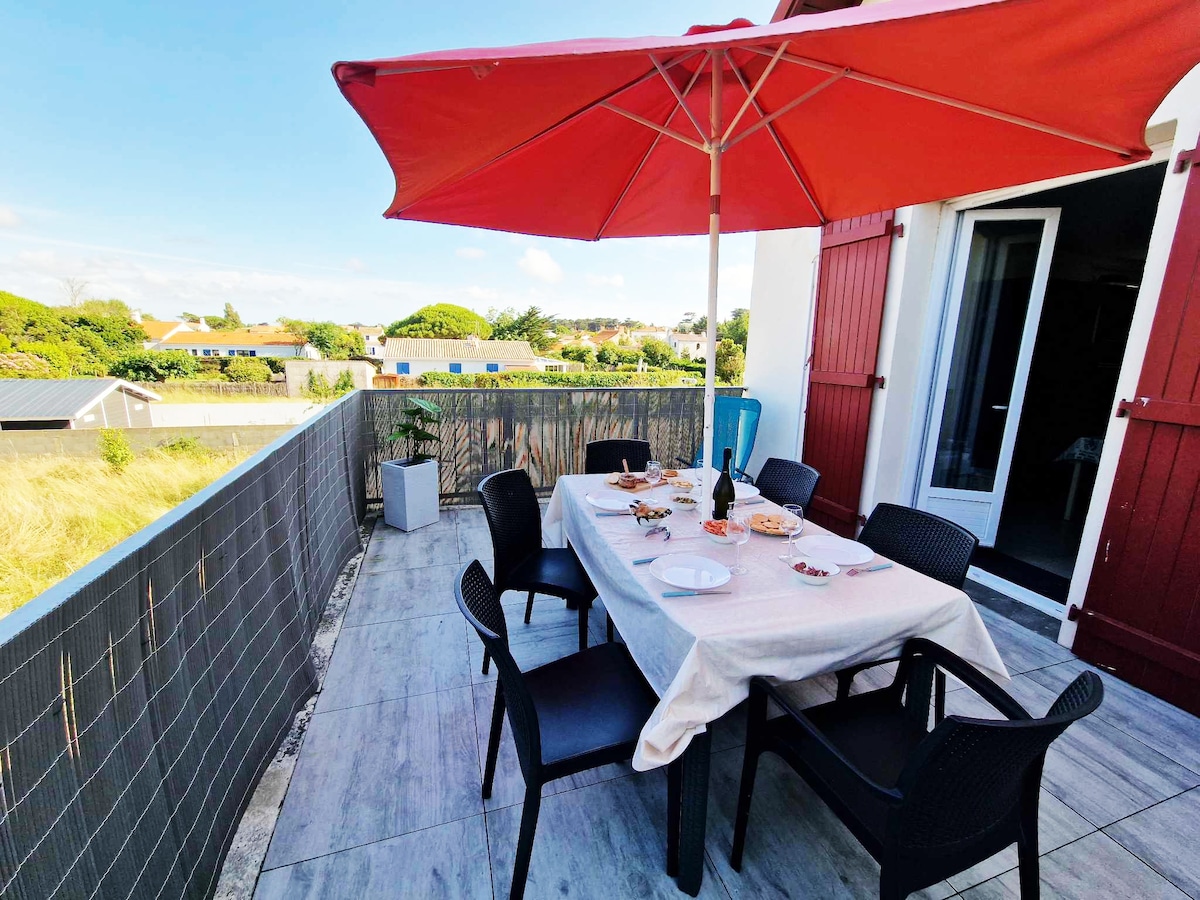 An outdoor terrace is presented with a large table covered in a light-colored tablecloth, surrounded by six black chairs. A red umbrella provides shade, and the surrounding area features greenery and distant rooftops. The terrace offers direct access to the living space.