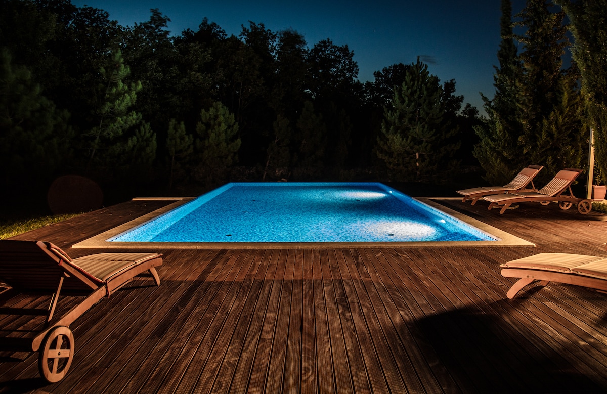 A private infinity pool is illuminated with soft blue lighting, surrounded by a wooden deck. Comfortable lounge chairs are positioned nearby, framed by lush greenery. The tranquil scene is set against a backdrop of dark trees, highlighting the peaceful ambiance of the outdoor space.