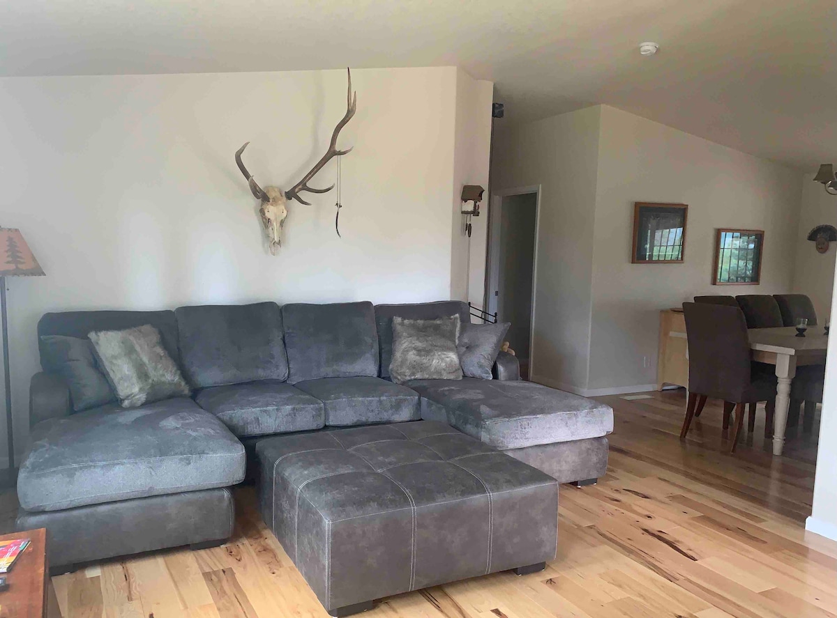 A spacious living area features a large sectional sofa with a plush texture facing a wall adorned with a mounted animal skull. A wooden coffee table sits in front, while a dining area can be seen in the background, accompanied by subtle wall decor.